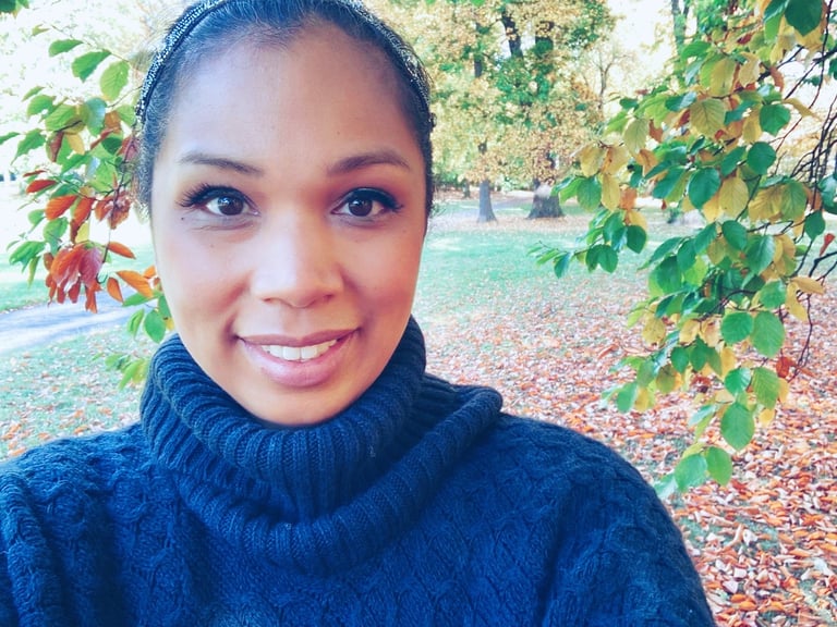 Woman smiling at camera in dark turtleneck sweater, outdoors among fall foliage and trees